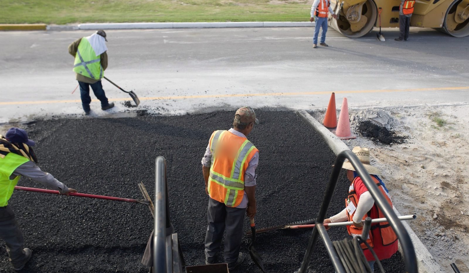 A crew in hi-vis vests rakes fresh hot mix across a new patch while the roller works the street beyond.