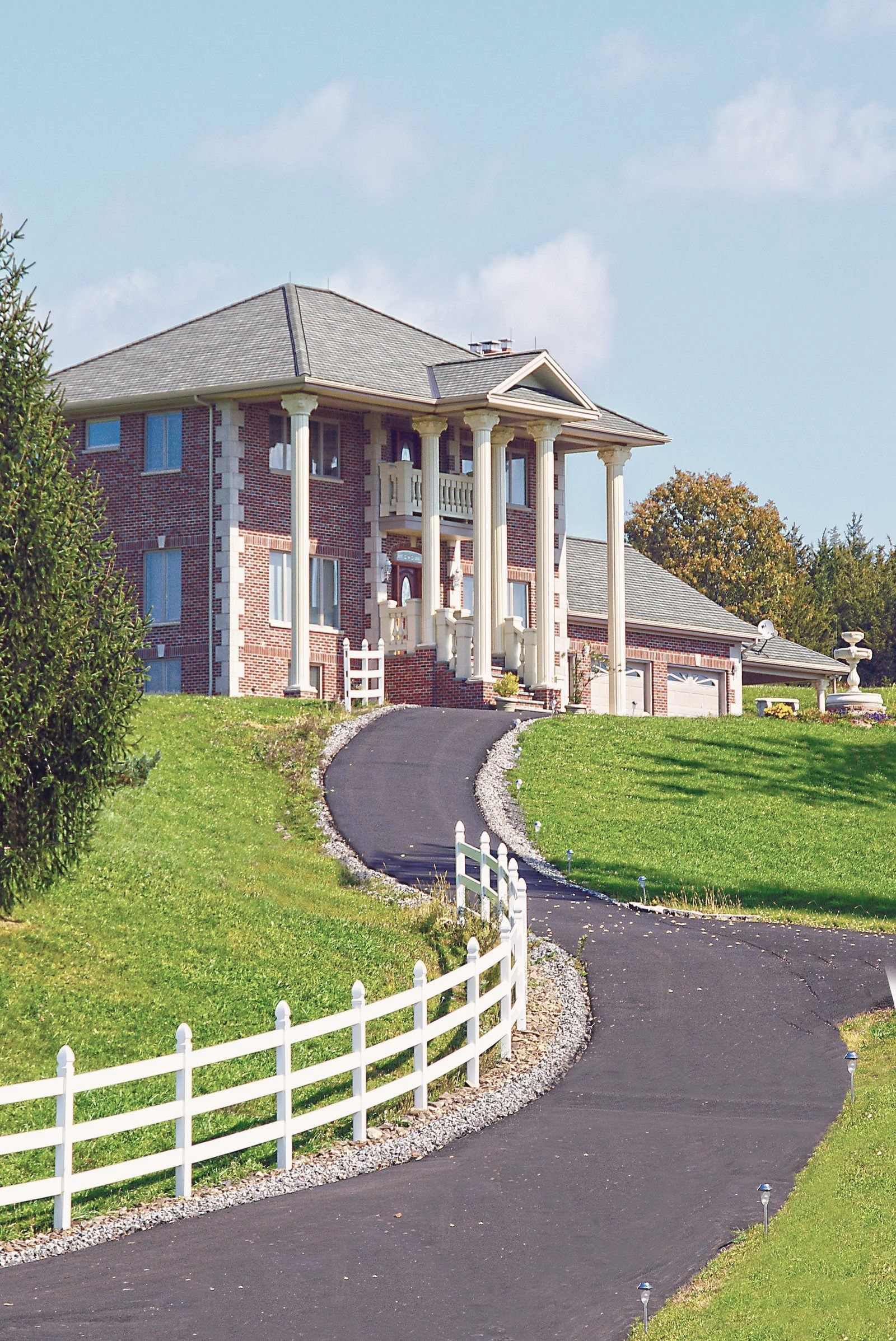 A smooth blacktop driveway curving up a fenced lawn to a brick house.