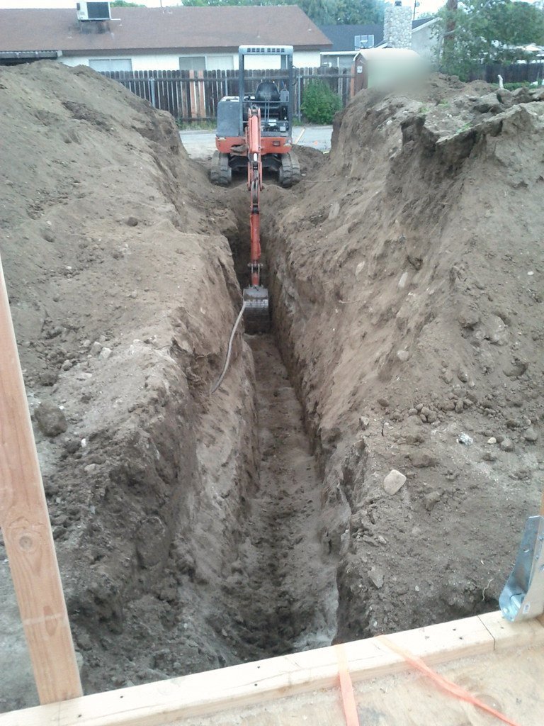 A freshly dug sewer trench runs through brown soil at a house lot, the mini excavator parked at its head.