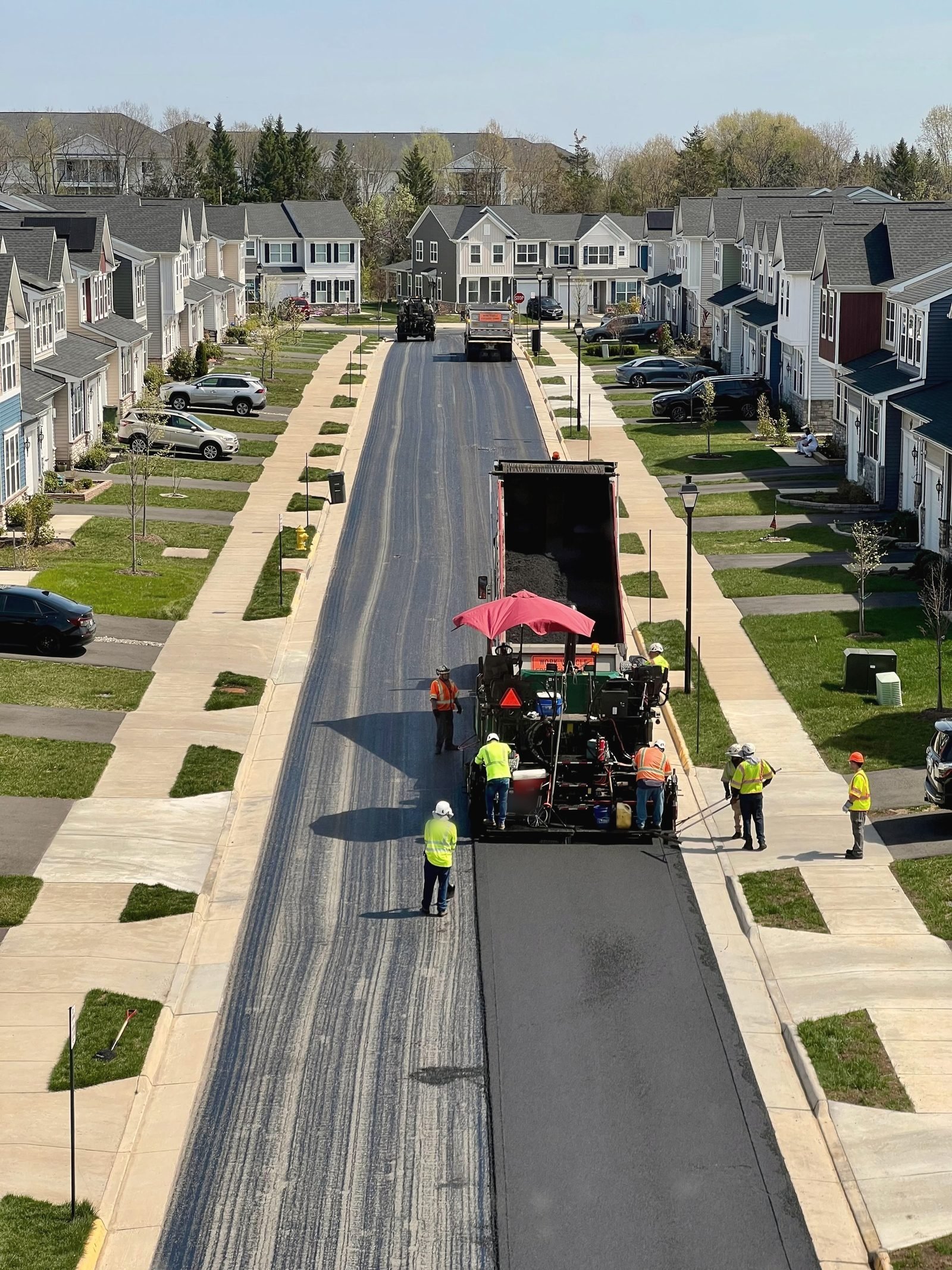 A paver and crew lay a new black surface down the middle of a residential street.